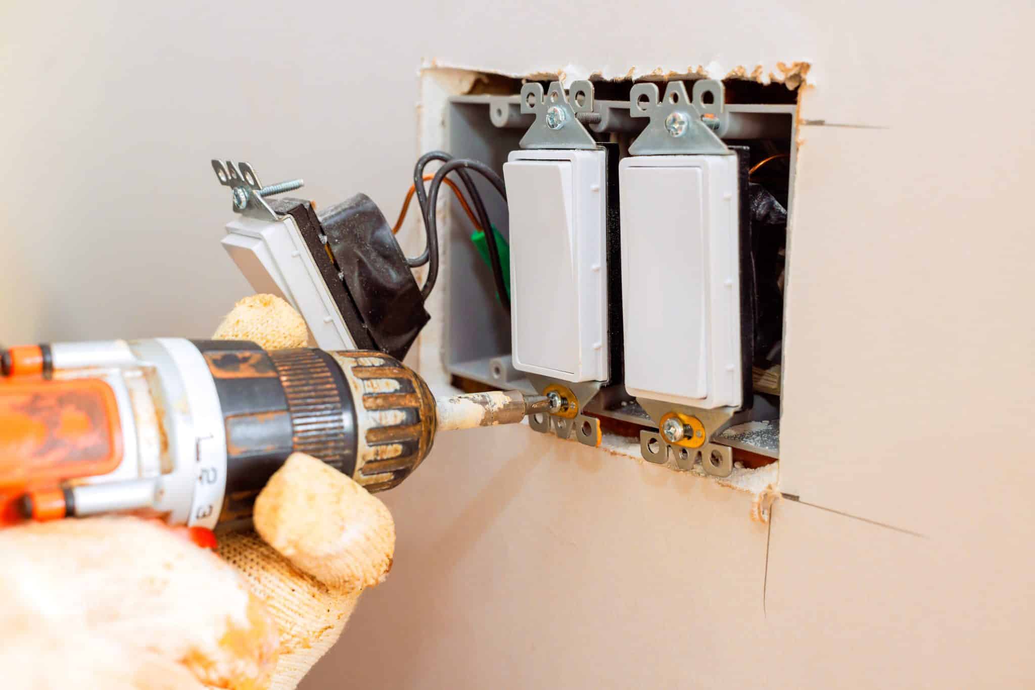 A pair of hands in worn white working gloves hold an electric drill to screw in a lightswitch panel.