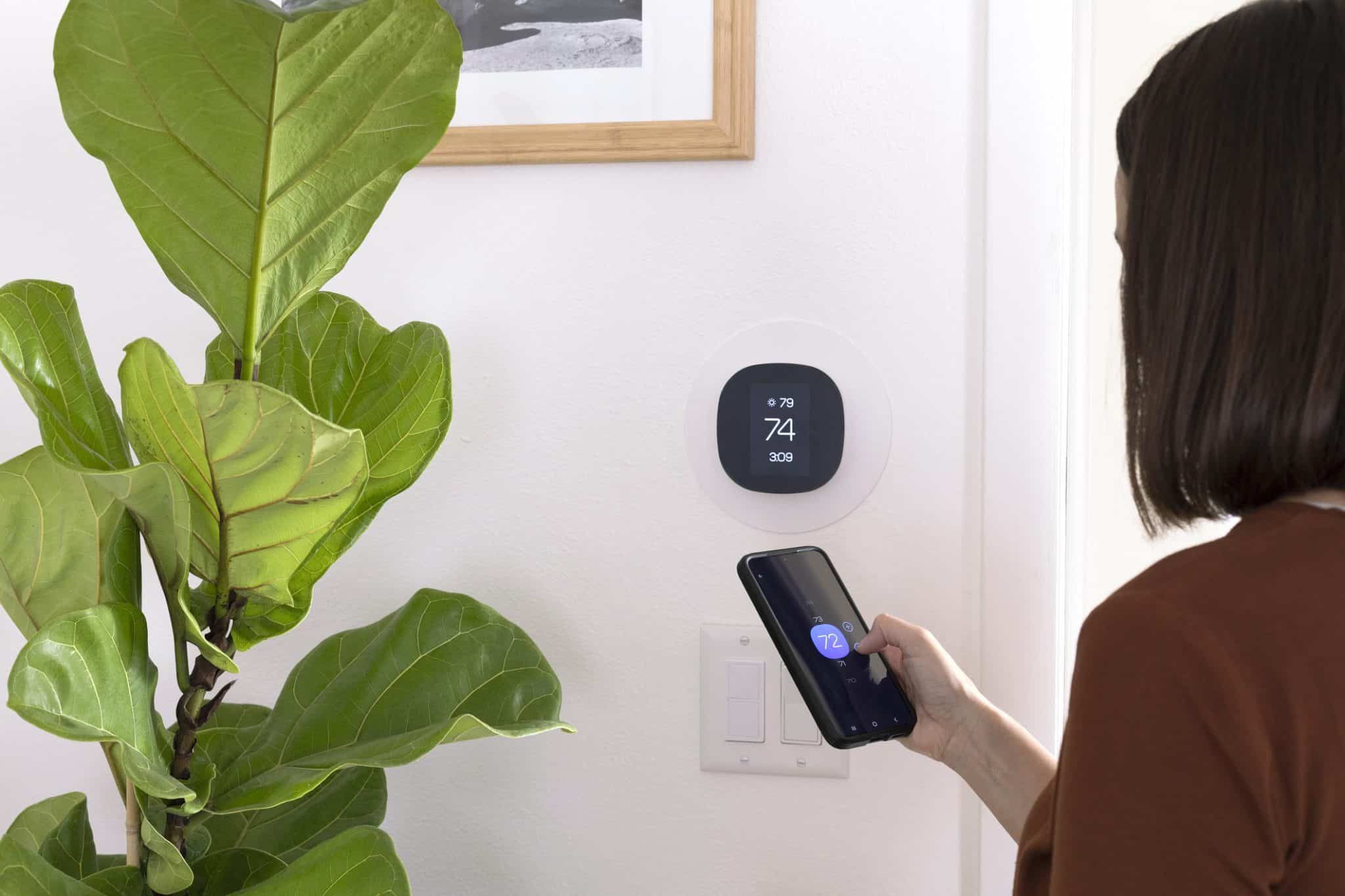 A homeowner in St. Charles adjusting a smart thermostat mounted on the wall, with a smartphone app open showing home temperature controls.