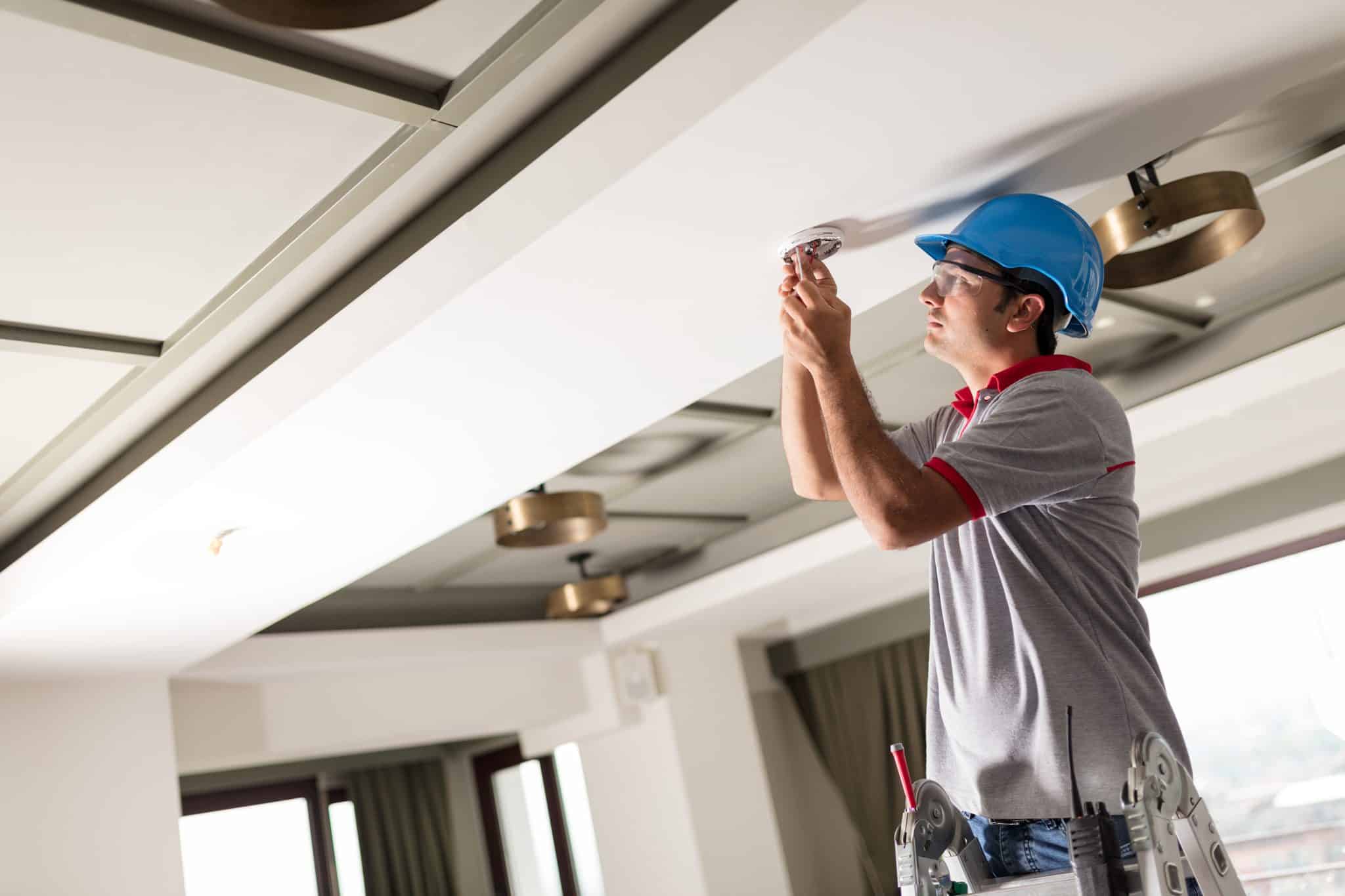 An electrician mounting a hardwired smoke and CO detector on a bedroom hallway ceiling in a St. Charles home.